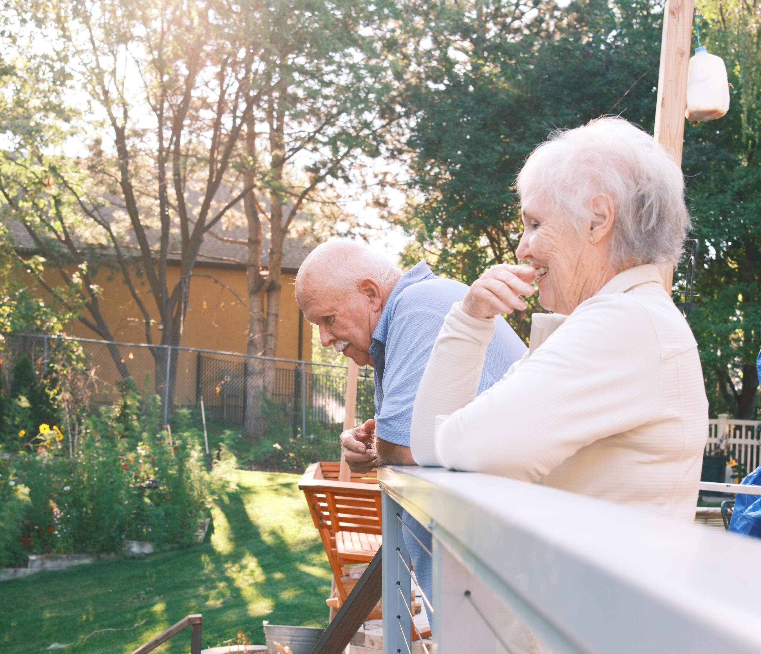An older couple laughing together on a sunny backyard deck surrounded by a lush garden – the inspiration behind Meadow Lane Tiles and the story of slow gatherings on Meadow Lane.
